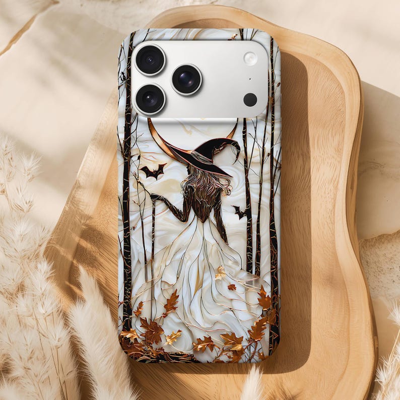 May include: A phone case with a witch design, wearing a black hat and white dress, set against a backdrop of bare trees and bats. The design incorporates gold and brown autumn leaves, creating a Halloween or autumn theme.