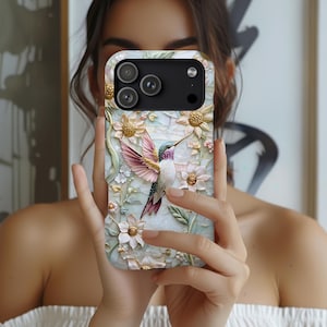 May include: A smartphone case featuring a raised, textured design of a hummingbird in flight surrounded by pale pink and white flowers with gold centres. The background has a marbled, off-white appearance with soft green leaves and stems.