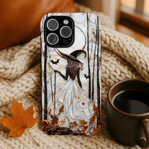 May include: A phone case with a witch design. The case has a white background with a witch in a black hat, a crescent moon, bats, and autumn leaves. The case is on a cream-coloured knitted blanket.