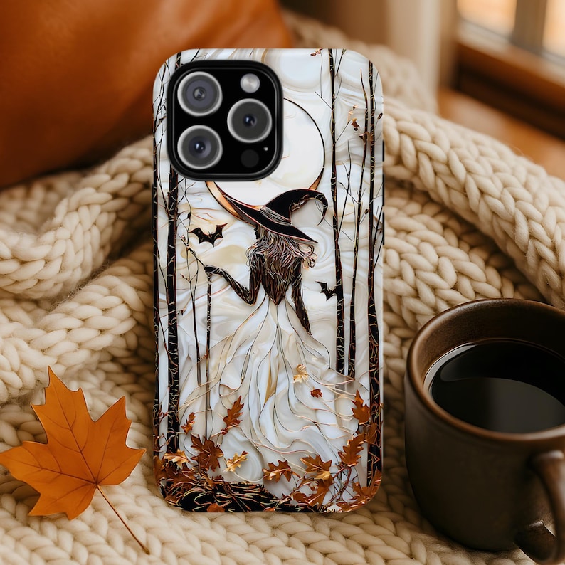 May include: A phone case with a witch design. The case has a white background with a witch in a black hat, a crescent moon, bats, and autumn leaves. The case is on a cream-coloured knitted blanket.