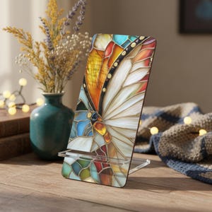 May include: A decorative phone stand featuring a colorful stained-glass butterfly design. The stand has a clear acrylic base and a vibrant display of orange, yellow, blue, and white hues. The butterfly's wings are intricately detailed, adding a touch of elegance.