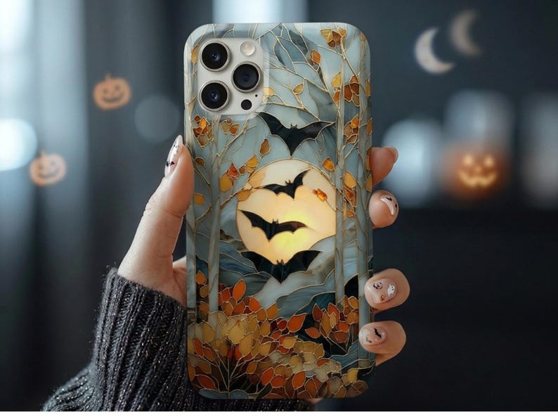 May include: A smartphone case featuring a stained-glass style design with a large yellow full moon, black silhouettes of flying bats, and autumn-colored foliage in shades of orange, brown, and gold against a pale blue and grey background.