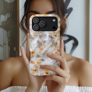 Puede incluir: Una funda para smartphone con fondo blanco decorada con peque&ntilde;as flores amarillas y azules y dos figuras de fantasmas blancos. La funda tiene un recorte negro para el m&oacute;dulo de c&aacute;mara en la parte superior con tres lentes.