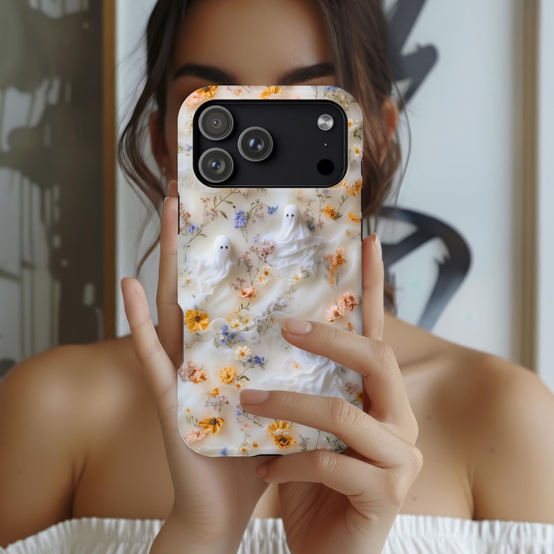 Puede incluir: Una funda para smartphone con fondo blanco decorada con peque&ntilde;as flores amarillas y azules y dos figuras de fantasmas blancos. La funda tiene un recorte negro para el m&oacute;dulo de c&aacute;mara en la parte superior con tres lentes.