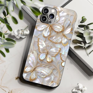 Peut inclure: Coque de téléphone blanche nacrée ornée de papillons aux contours dorés. Le motif présente des ailes de papillon détaillées sur un fond blanc avec de subtils motifs floraux. La coque a une finition brillante.