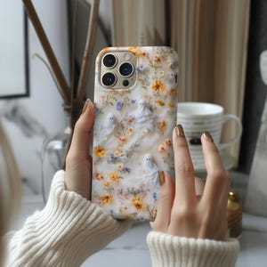 Puede incluir: Una funda de teléfono blanca con un diseño floral y pequeñas figuras de fantasmas. La funda presenta flores amarillas, naranjas y azules. La funda del teléfono la sostiene una persona que lleva un suéter color crema.