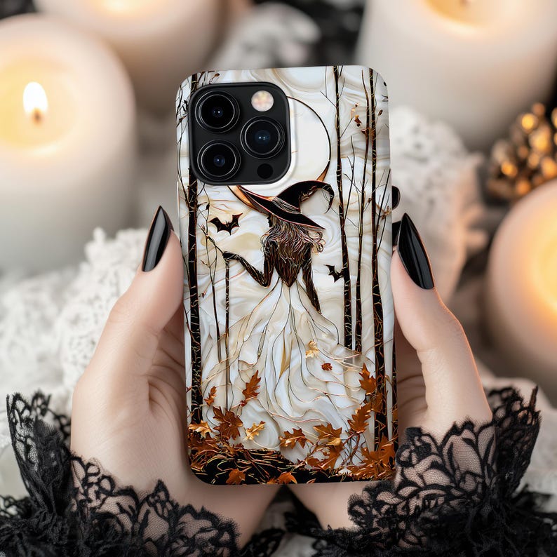 May include: A phone case with a witch illustration in a white dress and black hat, set against a backdrop of trees, a full moon, and bats. The case has a white and gold colour scheme with autumn leaves.