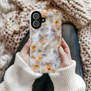 May include: A phone case with a cream-colored background featuring three white ghost figures and scattered small yellow, orange, and blue dried flowers. The case has a black camera cutout section at the top left corner with two circular lenses.