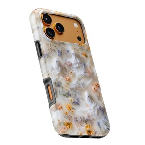 Puede incluir: Una funda para tel&eacute;fono con fondo color crema y un patr&oacute;n de peque&ntilde;as flores prensadas en tonos amarillos, naranjas y azules, intercaladas con tres figuras de fantasmas blancos. El &aacute;rea de la c&aacute;mara es de color dorado met&aacute;lico s&oacute;lido.
