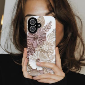 May include: A white smartphone case featuring a textured, three-dimensional floral design. The pattern includes various layered flowers and fern-like leaves in shades of dusty rose, cream, and beige, with a prominent dual-lens camera cutout.