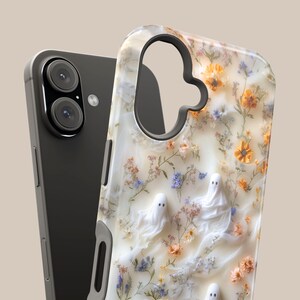 Puede incluir: Una funda para tel&eacute;fono con fondo color crema, decorada con un patr&oacute;n de peque&ntilde;as flores silvestres naranjas y azules y tres figuras de fantasmas blancos. La funda tiene un gran recorte para la lente de la c&aacute;mara y un smartphone gris oscuro.