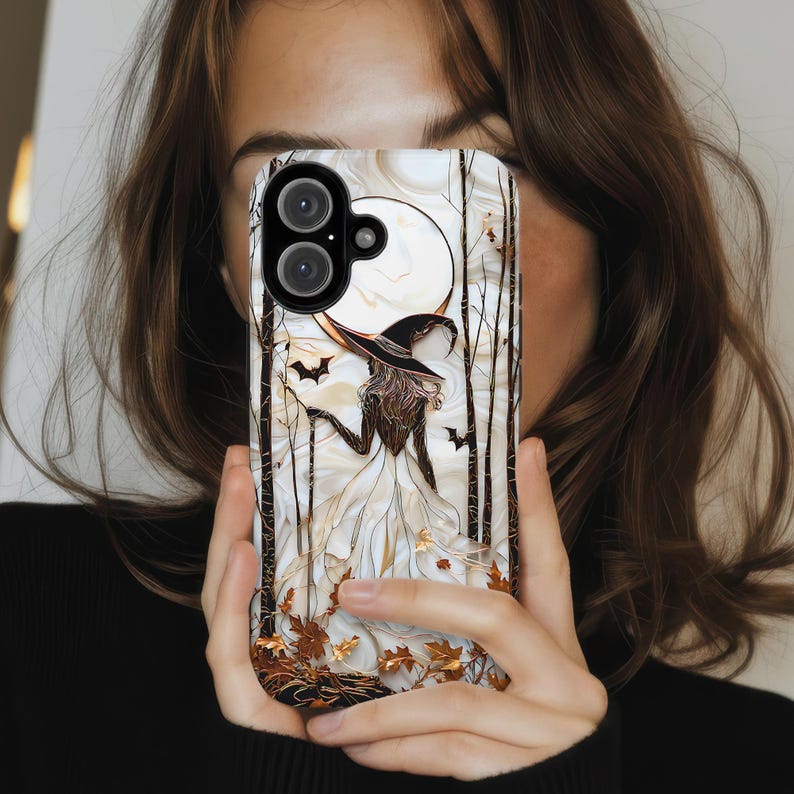 May include: A phone case with a witch illustration, a full moon, bats, and autumnal leaves. The witch is depicted in a black hat and white dress. The background has a marble effect with brown and gold detailing.