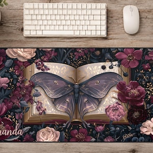 May include: A rectangular desk mat featuring an open book with a large, dark purple moth resting on its pages, surrounded by a dense arrangement of deep red, pink, and dark purple flowers and foliage, with the name Amanda written in white script.