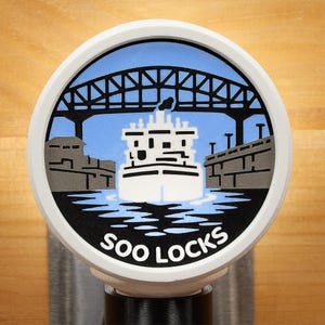 May include: A round, white and black tap handle featuring a blue sky and a black bridge over a white ship. The words "SOO LOCKS" are printed in white on the bottom of the handle.