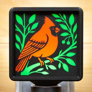 May include: A square, black night light featuring a vibrant orange cardinal bird design. The bird is perched on a branch with green leaves and berries against a black background. The night light is mounted on a silver base.