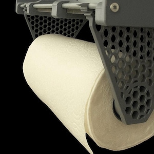 Könnte beinhalten: Ein grauer Papierhandtuchhalter mit einer Rolle weißer Papierhandtücher. Der Halter hat ein geometrisches Design mit sechseckigen Aussparungen. Die Papierhandtücher sind strukturiert und wirken weich. Der Hintergrund ist schwarz.