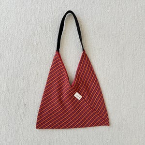 Puede incluir: Un bolso tote a cuadros rojos y amarillos con una correa negra. El bolso tiene una forma triangular &uacute;nica y una peque&ntilde;a etiqueta blanca en la parte delantera. El bolso est&aacute; hecho de tela tejida.