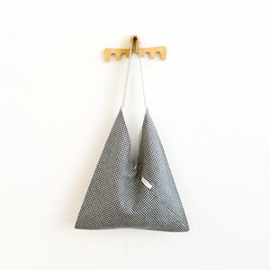 May include: A triangular-shaped tote bag featuring a black and white gingham check pattern. The bag has a single integrated shoulder strap and a small white rectangular fabric label attached to the front side near the opening of the bag.