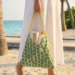 May include: A green and white patterned beach bag with yellow straps is held by a person wearing a white dress. The bag contains a rolled towel and a bottle of sunscreen. The bag is on a sandy beach.