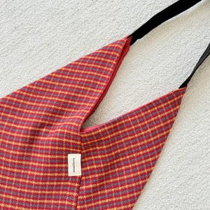 Puede incluir: Bolso de hombro a cuadros rojos y amarillos con una correa negra. El bolso tiene una forma triangular &uacute;nica y una peque&ntilde;a etiqueta blanca con el texto "lovingyana". El bolso est&aacute; hecho de tela tejida.