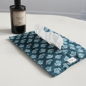 May include: Teal fabric tissue box cover with a white leaf pattern. White tissues protrude from the opening. A dark brown bottle with a label reading "Silent Flame" is in the background. The tissue box cover has a small white tag that reads "lovinggreen".