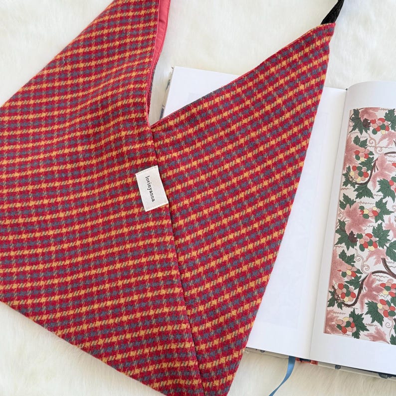 Puede incluir: Un bolso tote con estampado rojo, amarillo y gris con una correa negra. El bolso tiene forma triangular y una peque&ntilde;a etiqueta blanca con texto. Se ve un libro abierto con ilustraciones florales.