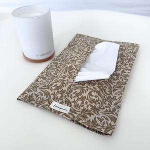 May include: A white cylindrical candle holder with a wooden base and the text Brightfield, alongside a rectangular fabric tissue pouch featuring a brown and beige swirling leaf pattern and a small white label with the text lovinganna.