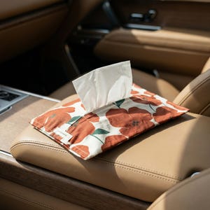 Puede incluir: Un dispensador de pañuelos rectangular con un estampado floral de flores color óxido y hojas verdes sobre fondo blanco. Se ven pañuelos blancos saliendo. El dispensador está sobre una superficie de cuero color beige.