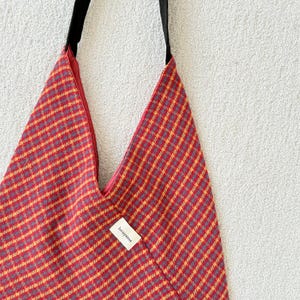 Puede incluir: Un bolso tote con estampado rojo y amarillo con una correa negra. El bolso tiene una forma triangular &uacute;nica y una peque&ntilde;a etiqueta que dice "loriyanna". El interior de la bolsa es rojo.