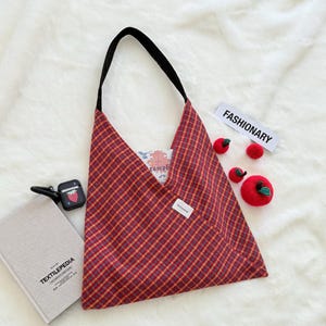 Puede incluir: Un bolso tote a cuadros rojos y dorados con una correa negra. El bolso tiene un bolsillo y contiene un libro y otros art&iacute;culos. La palabra "FASHIONARY" est&aacute; en una etiqueta blanca. Tambi&eacute;n hay varios art&iacute;culos de ganchillo con forma de manzana roja.
