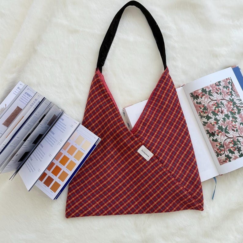 Puede incluir: Un bolso tote a cuadros rojos y dorados con una correa negra. El bolso tiene un dise&ntilde;o en forma de V y una peque&ntilde;a etiqueta blanca. Libros abiertos y muestras de color son visibles cerca del bolso.