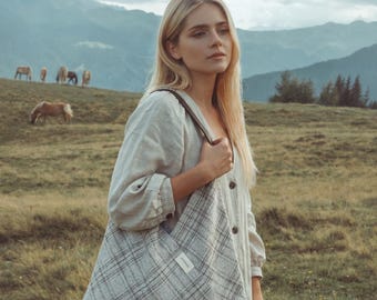 May include: A woman holding a large, plaid tote bag with brown straps. The bag is gray and white with a grid pattern. The woman is wearing a light-colored dress and standing in a grassy field with mountains in the background.