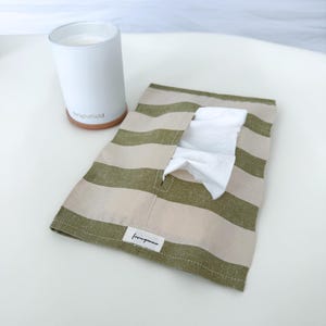 Könnte beinhalten: Ein rechteckiger Stoff-Taschentuchhalter mit horizontalen olivgrünen und beigen Streifen und einer mittigen Öffnung für weiße Taschentücher. Ein kleines weißes Etikett mit dem schwarzen Text "lovinganna" ist am unteren Rand angenäht.