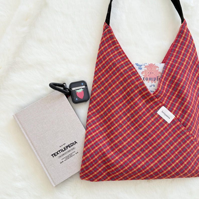 Puede incluir: Un bolso tote a cuadros rojos y amarillos con una correa negra, un libro titulado "TEXTILEPEDIA" y una funda negra para AirPod con un dise&ntilde;o de fresa. El bolso tiene un bolsillo con un estampado floral en el interior.