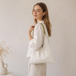 May include: A white long-sleeved button-down shirt paired with a triangular-shaped tote bag featuring a white and beige striped pattern and a solid olive green shoulder strap. The bag is worn over the shoulder, showcasing its unique geometric design.