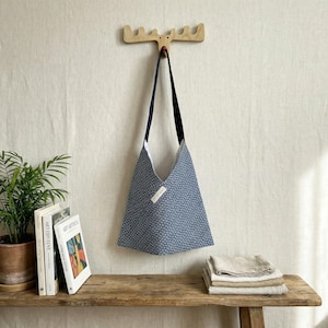 Puede incluir: Un bolso tote con estampado azul y blanco con una correa negra cuelga de un gancho de pared con forma de cabeza de alce de madera. La bolsa es triangular y tiene un interior blanco. Una pequeña etiqueta blanca está adjunta en la parte delantera.