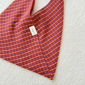Puede incluir: Un bolso tote a cuadros rojos y dorados con interior rojo. El bolso tiene una forma triangular &uacute;nica y una peque&ntilde;a etiqueta que dice "lorinyanna". El bolso est&aacute; hecho de una tela texturizada.