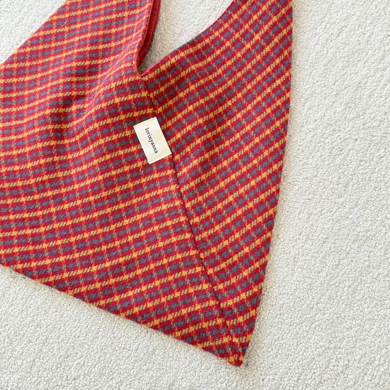 Puede incluir: Un bolso tote a cuadros rojos y dorados con interior rojo. El bolso tiene una forma triangular &uacute;nica y una peque&ntilde;a etiqueta que dice "lorinyanna". El bolso est&aacute; hecho de una tela texturizada.