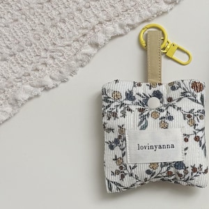 Op de afbeelding: Een vierkant, wit corduroy tasje met een bloemenpatroon in blauwe, bruine en gele tinten. Het tasje heeft een beige stoffen lus bevestigd aan een felgele plastic karabijnhaak. Een wit label met de tekst lovinyanna is in het midden geplaatst.