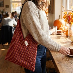 Puede incluir: Un bolso tote a cuadros burdeos y dorados con una correa negra. El bolso se lleva al hombro. La imagen est&aacute; tomada en interiores, con una persona con un su&eacute;ter beige y vaqueros azules. El bolso tiene una peque&ntilde;a etiqueta blanca.