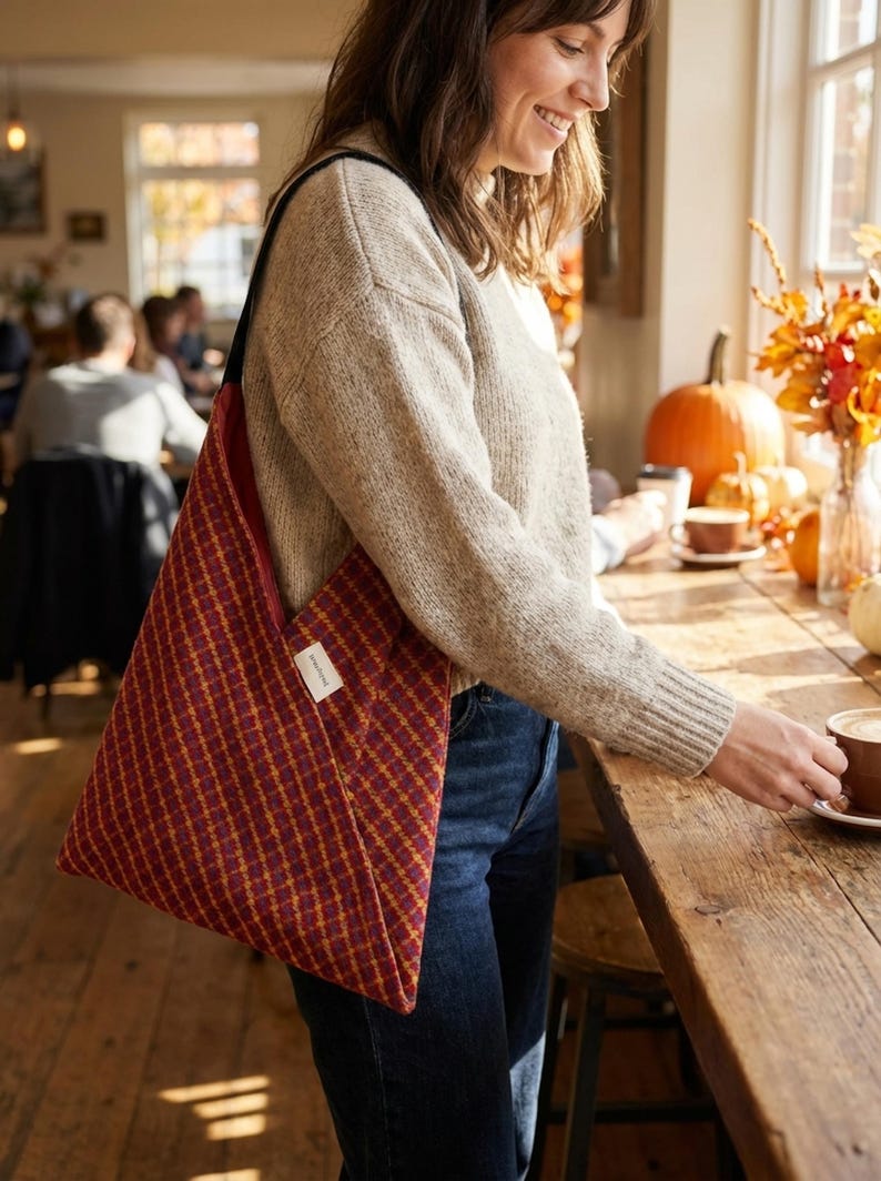 Puede incluir: Un bolso tote a cuadros burdeos y dorados con una correa negra. El bolso se lleva al hombro. La imagen est&aacute; tomada en interiores, con una persona con un su&eacute;ter beige y vaqueros azules. El bolso tiene una peque&ntilde;a etiqueta blanca.