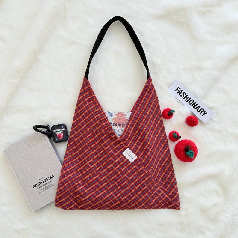 Puede incluir: Un bolso tote a cuadros rojos y amarillos con una correa negra, un libro y auriculares inal&aacute;mbricos. La bolsa tiene una forma triangular &uacute;nica. La palabra "Fashionary" est&aacute; impresa en una tarjeta blanca. Tambi&eacute;n hay manzanas de ganchillo.