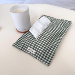 May include: A white candle in a matte container with the text Brightfield printed on the side, positioned next to a rectangular fabric tissue holder featuring a dark green and white gingham check pattern and a small label reading lovingyanna.