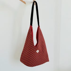 Puede incluir: Un bolso tote a cuadros rojos y amarillos con una correa negra cuelga contra una pared blanca. El bolso tiene forma triangular y un interior rojo. Una peque&ntilde;a etiqueta blanca est&aacute; cosida en la parte delantera.