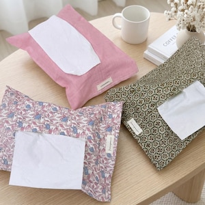 Puede incluir: Una variedad de fundas para cajas de pañuelos en varios patrones y colores. Una es rosa, otra tiene un diseño floral y la tercera es verde con un patrón geométrico. Cada funda contiene una pila de pañuelos blancos y está colocada sobre una mesa de madera.