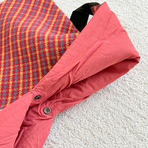 Puede incluir: Primer plano de un bolso tote. El exterior presenta un estampado de cuadros rojos y amarillos, mientras que el interior es de color coral liso. Tiene un asa negra y un cierre de bot&oacute;n a presi&oacute;n.