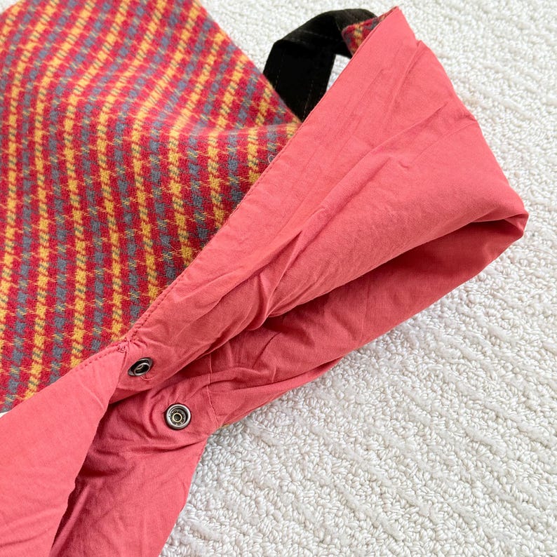 Puede incluir: Primer plano de un bolso tote. El exterior presenta un estampado de cuadros rojos y amarillos, mientras que el interior es de color coral liso. Tiene un asa negra y un cierre de bot&oacute;n a presi&oacute;n.