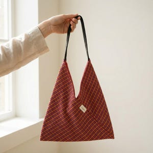 Puede incluir: Un bolso tote triangular con un patr&oacute;n de cuadros rojos y amarillos. Tiene correas negras y una peque&ntilde;a etiqueta blanca. La forma &uacute;nica del bolso se destaca al ser sostenido por una mano.