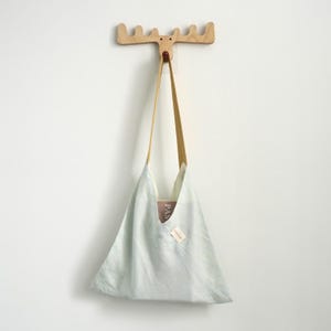 May include: A light blue tote bag with a long, thin strap hangs from a wooden moose head wall hook. The bag has a triangular shape and a light-colored interior. A small tag is attached to the bag.