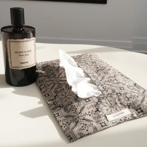 May include: A dark brown glass bottle with a black cap and a label that reads "SILENT FLAME Home Spray". Next to it is a beige tissue holder with a black floral pattern, and white tissues are visible. The tissue holder has a small tag that reads "LOVINYANNA".